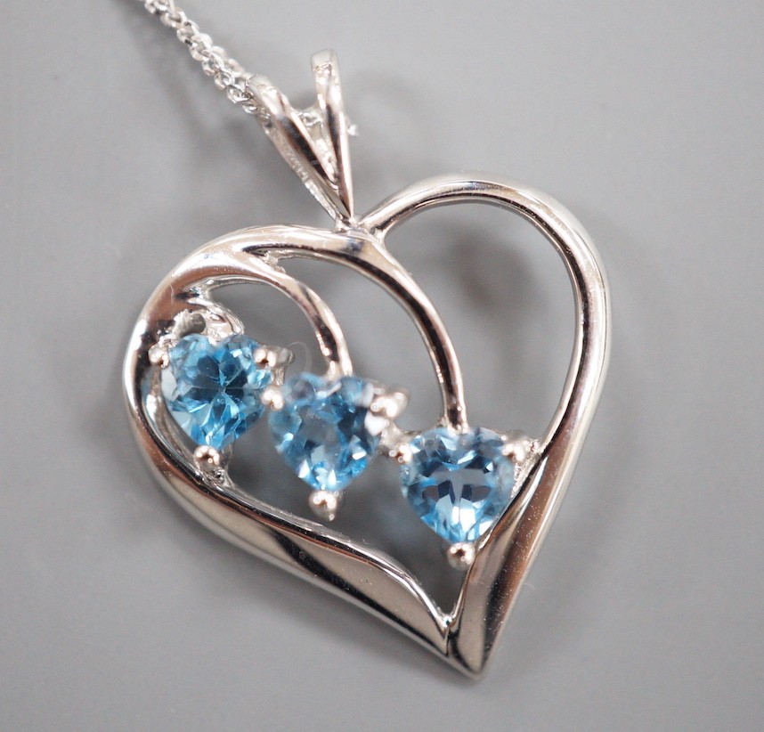 A modern 9ct white gold and three stone blue topaz set stylised heart shaped, pendant, 25mm, on a 375 fine link chain, 40cm, gross weight 2.7 grams.